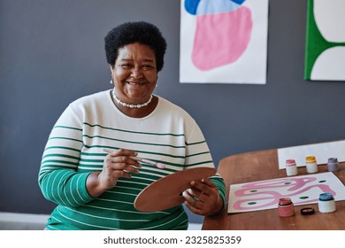 Successful mature teacher of painting with watercolor palette looking at you with smile while sitting by workplace in front of camera in studio - Powered by Shutterstock