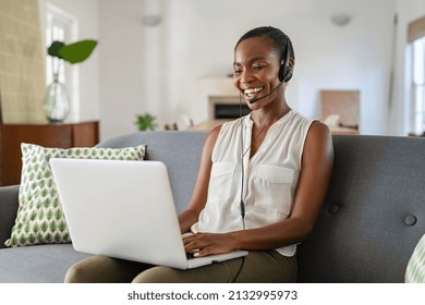 Successful Middle Aged African Woman With Headphone And Mic Talking On Video Call Using Laptop. Cheerful Mid Black Woman Enjoy Video Call On Laptop While Sitting On Couch Working From Home.