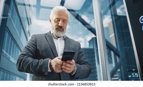 Successful Middle Aged Businessman Riding Glass Elevator To Office In Modern Business Center. Handsome Happy Man Using Smartphone, Write A Message, Social Media And Work Emails In A Lift.