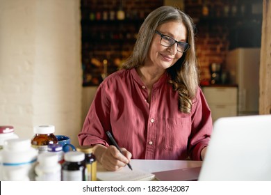 Successful Middle Aged Female Dietitian In Rectangular Glasses Working From Home Having Online Consultation With Patient Via Video Conference Call On Laptop Computer, Handwriting, Making Prescription