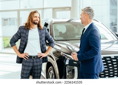 Successful middle-aged businessman in a business suit is selling cars to a happy smiling handsome male buyer in a car showroom with crossover on background. - Shutterstock ID 2236141535