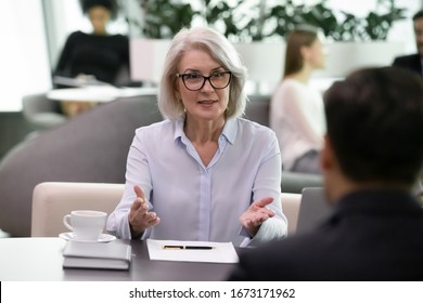 Successful Middle-aged Businesswoman Sit At Desk Talk With Business Colleague At Meeting, Confident Senior Female Boss Lead Interview With Job Candidate Applicant In Office, Employment Concept