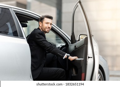 Successful middle-aged entrepreneur getting in expensive car, driving to airport, side view. Smiling bearded businessman sitting in car, going to business meeting or conference, empty space