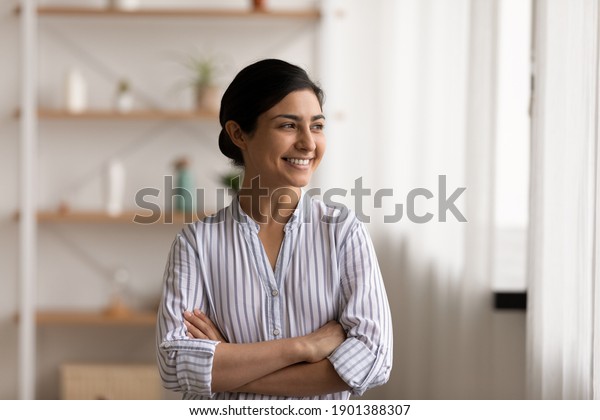 Successful millennial indian lady stand at home\
keep arms crossed on chest look at window with confident smile.\
Happy mixed race female private enterpreneur proud of being self\
made business woman
