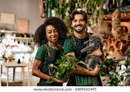 A successful multinational smiling family of small business owners of a plant shop. The woman is holding a flower pot, the man is jokingly holding a gray British cat.