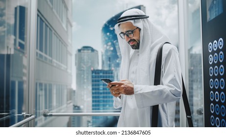 Successful Muslim Businessman In Traditional White Kandura Riding Glass Elevator To Office In Modern Business Center. Man Using Smartphone. Saudi, Emirati, Arab Businessman Concept.