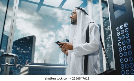 Successful Muslim Businessman In Traditional White Kandura Riding Glass Elevator To Office In Modern Business Center. Man Using Smartphone. Saudi, Emirati, Arab Businessman Concept.