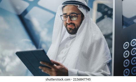 Successful Muslim Businessman In Traditional White Kandura Riding Glass Elevator To Office In Modern Business Center. Man Using Tablet Computer. Saudi, Emirati, Arab Businessman Concept