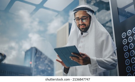 Successful Muslim Businessman In Traditional White Kandura Riding Glass Elevator To Office In Modern Business Center. Man Using Tablet Computer. Saudi, Emirati, Arab Businessman Concept.