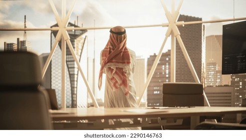 Successful Muslim Businessman in Traditional White Outfit Standing in His Modern Office Looking out of the Window on Big City with Skyscrapers. Successful Saudi, Emirati, Arab Businessman Concept