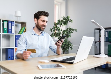 Successful online shopping through a smartphone. Successful deal. Satisfied young man holds a credit card and a mobile phone, makes an order. Sitting at a desk in the office on a laptop