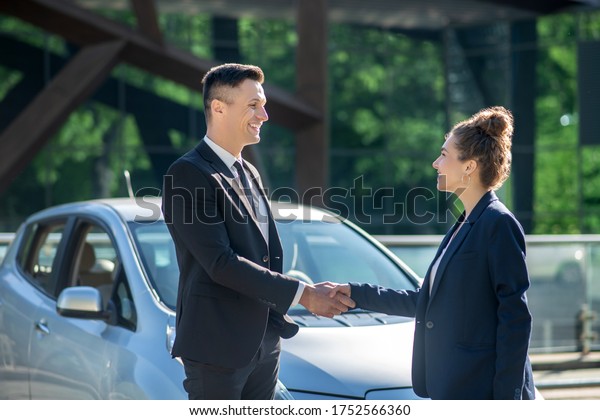 Successful people. Young successful man\
and woman shaking hands near a car, looking\
joyful