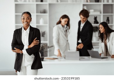 Successful person. Confident african american businesswoman posing with folded arms and smiling at camera in office. Diverse colleagues working on background - Shutterstock ID 2353199185