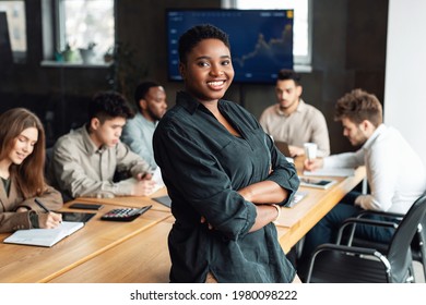 Successful Person. Portrait Of Confident Smiling African American Businesswoman Sitting Leaning On Desk In Office, Posing With Folded Arms And Looking At Camera, Colleagues Working In Background