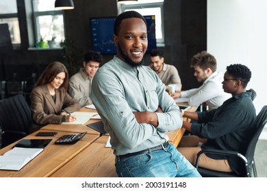 Successful Person. Portrait Of Confident Smiling African American Businessman Sitting Leaning On Desk In Office, Posing With Folded Arms And Looking At Camera, Diverse Colleagues Working In Background