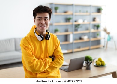 Successful Person. Portrait Of Confident Smiling Asian Man In Bright Yellow Hoodie And Headset Sitting Leaning On Desk, Posing With Folded Arms And Looking At Camera. Home Office In Blurred Background