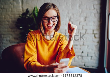 Successful positive young woman with short haircut watching live stream online on smartphone and celebrating victory of favourite team.Emotional hipster girl happy with win in online contest