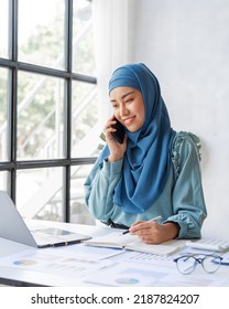 Successful Pretty Asian Muslim Lady In Hijab Make A Phone Call When Working At Desk In Office During Working On Her Project On Laptop Computer.