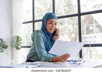 Successful Pretty Asian Muslim Lady In Hijab Make A Phone Call When Working At Desk In Office During Working On Her Project On Laptop Computer.