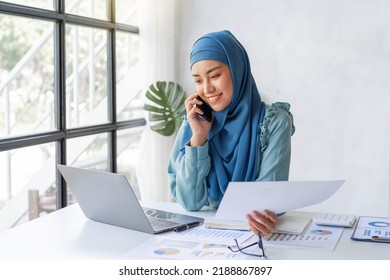 Successful Pretty Asian Muslim Lady In Hijab Make A Phone Call When Working At Desk In Office During Working On Her Project On Laptop Computer.