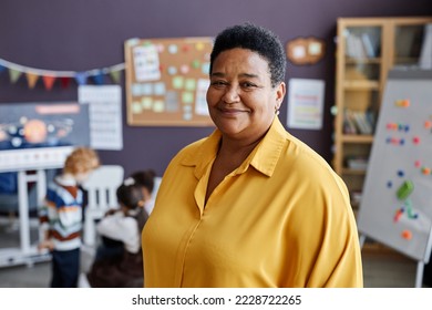 Successful professional mature female teacher of nursery school looking at camera while standing against group of little learners - Powered by Shutterstock