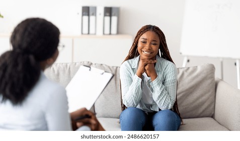 Successful psychotherapy. Millennial female client having session with psychologist, smiling, sitting on couch at clinic. Cheerful black lady communicating with therapist, solving emotional problem - Powered by Shutterstock