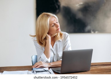 Successful relaxed tired middle aged caucasian blonde woman, business lady or lawyer, wearing white shirt, sitting at a table, taking break, closes eyes, overworked, exhausted