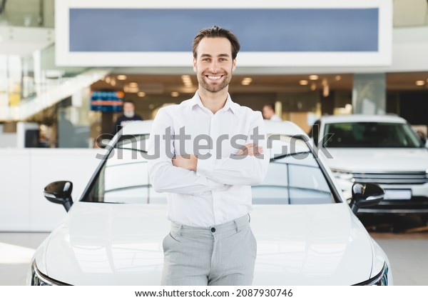 Successful rich man in formal\
clothes buying purchasing new expensive car automobile in auto\
trader shop store dealer, leaning on the car with his arms\
crossed