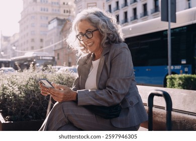 Una exitosa profesional senior disfruta de un descanso, mirando a su App de smartphone en un banco de la ciudad.El fondo muestra un autobús, que representa los viajes, la movilidad empresarial y un entorno de trabajo urbano dinámico