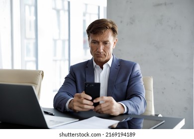 Successful serious middle-aged businessman using smartphone seated at workplace in skyscrapers office. Modern tech, business application usage, corporate messaging, agenda, appointments memo concept