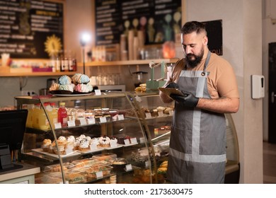 Successful Small Business Owner In Cafe Using Digital Tablet And Looking At Screen. Serious Barista With A Beard Standing In Front Of Showcase While Counting, Start Up Coffee Shop