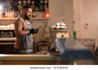 Successful small business owner in cafe using digital tablet and looking at screen. Serious barista with a beard standing in front of showcase while counting, SME concept - Powered by Shutterstock