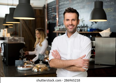 Successful small business owner standing with crossed arms with employee in background preparing coffee - Shutterstock ID 286965218