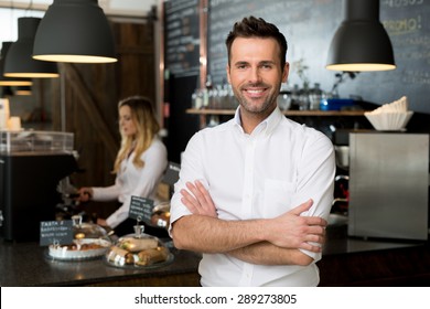 Successful small business owner standing with crossed arms with employee in background preparing coffee