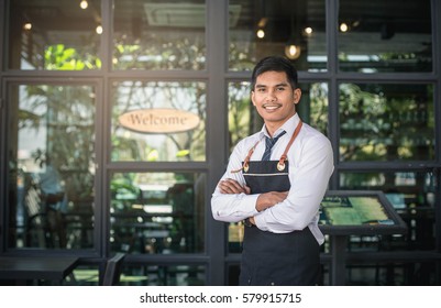 Successful Small Business Owner Standing With Crossed Arms With Employee In Background Coffee Shop.