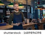 Successful small business owner using digital tablet and looking at camera. Happy smiling waiter ready to take order. Portrait of young entrepreneur of coffee shop standing at counter with copy space.