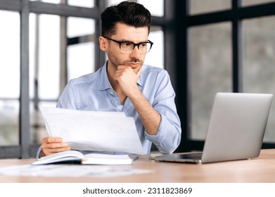 Successful smart focused young caucasian business man, freelancer or manager sitting at the table in the office, looking through documents, thinking over business strategy, looking at laptop screen - Shutterstock ID 2311823869