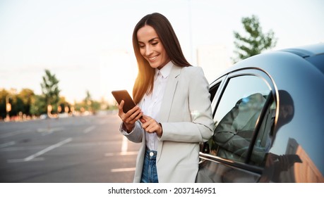 Successful smiling attractive woman in formal smart wear is using her smart phone while standing near modern car outdoors - Powered by Shutterstock