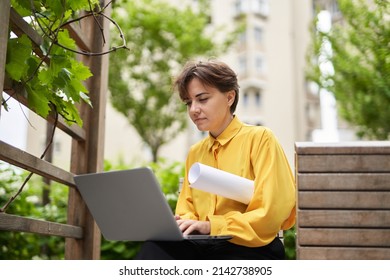 Successful Smiling Business Lady In Yellow Blouse Sitting On Bench Outdoor In Park With Plans Using Laptop Working Or Having Online Meeting. Business, Finance Industry Or Real Estate Concept