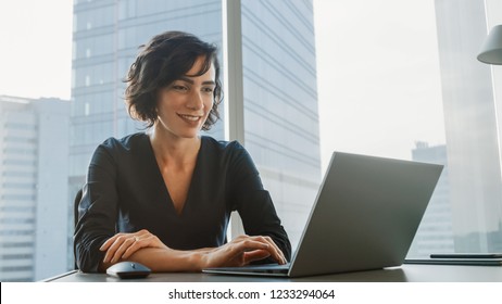 Successful Smiling Businesswoman Working on a Laptop in Her Modern Office. Stylish Beautiful Woman Doing Important Job. In the Window Big City Business District View.