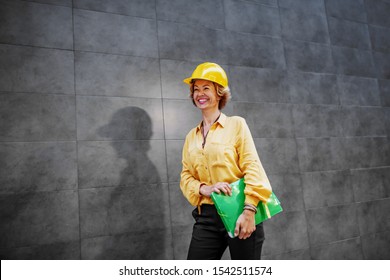 Successful Smiling Caucasian Blonde Senior Female Architect With Helmet On Head And Folder With Documents Under Armpit Passing By Gray Wall And Going To The Construction Site.