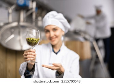 Successful smiling female brewer in white uniform showing natural granulated hops while standing in brewery. Selective focus on glass with pellets. Concept of high quality raw materials.. - Powered by Shutterstock