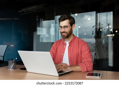 Successful smiling man working inside office with laptop, businessman in red shirt smiling and typing on keyboard in glasses, programmer working software for program. - Powered by Shutterstock