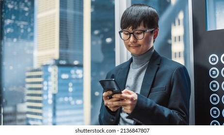 Successful Stylish Japanese Businessman Riding Glass Elevator To Office In Modern Business Center. Handsome Man Smile While Using Smartphone, Write Text Message, Check Social Media In A Lift.