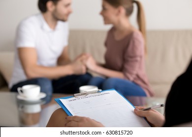 Successful therapy session with family couple in the background, reconciliation after argument, making peace, focus on hands of female psychologist holding clipboard with medical card, close up