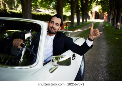 Successful And Wealthy Businessman Sitting Behind Steering Wheel Of Luxury Cabriolet Car On Countryside Road, Confident Handsome Man Sits In His New Convertible Car Doing Super Gesture With The Finger 