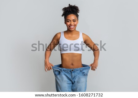 Successful weight loss, afro woman with too large jeans after effective diet, grey background