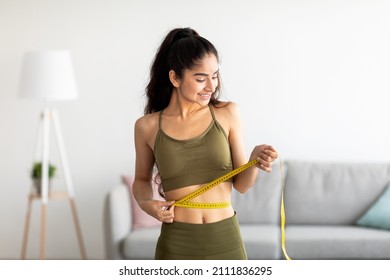 Successful weight loss concept. Fit young Indian woman in sports clothes measuring waist with tape at home. Millennial lady feeling satisfied with slimming program indoors