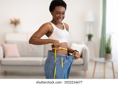Successful Weight Loss. Joyful Young Black Lady Measuring Waist Wearing Oversized Jeans After Slimming, Smiling To Camera Standing Indoors. Weight-Loss Comparison And Motivation