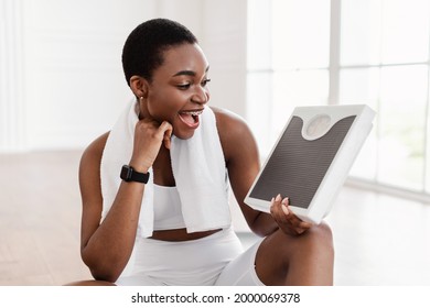 Successful Weight Loss. Portrait Of Excited Black Woman Holding And Looking At Scales Wearing White Sportswear And Towel After Workout. Happy Lady Achieved Her Weight-Loss Goal